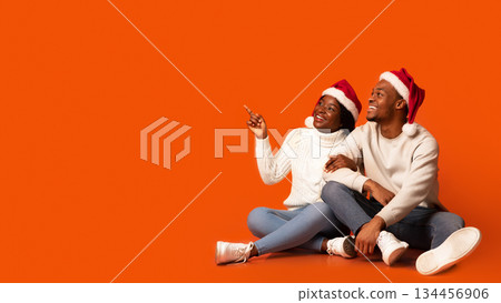 Happy black spouses wearing santa hats sitting on orange background, lady pointing away with finger, smiling african american couple demonstrating copy space for your christmas greetings or sale offer Happy black spouses wearing santa hats sitting on orange background, lady pointing away with finger, smiling african american couple demonstrating copy space for your christmas greetings or sale offer 134456906