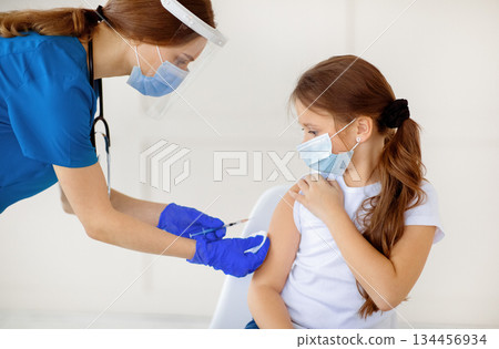 Covid-19 vaccination campaign. Little girl in protective mask receiving coronavirus vaccine injection from doctor at hospital. Antiviral immunization, infectious disease prevention concept Covid-19 vaccination campaign. Little girl in protective mask receiving coronavirus vaccine injection from doctor at hospital. Antiviral immunization, infectious disease prevention concept 134456934