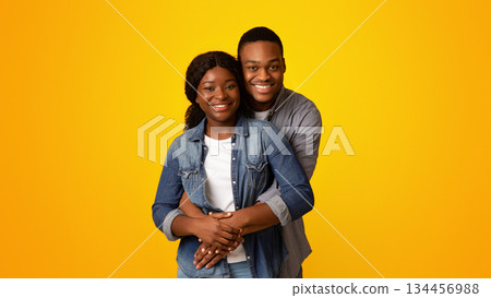Portrait of happy african american lovers hugging and smiling at camera while standing together isolated over yellow background, romantic black guy cuddling his girlfriend from back, copy space Portrait of happy african american lovers hugging and smiling at camera while standing together isolated over yellow background, romantic black guy cuddling his girlfriend from back, copy space 134456988