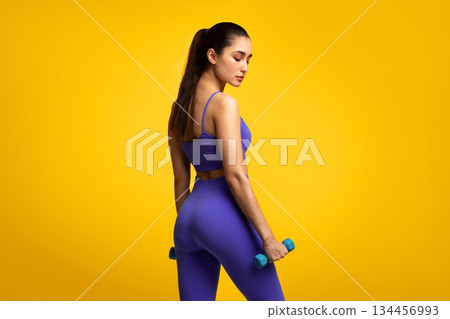 Fitness Model. Portrait Of Confident Young Woman Exercising With Two Dumbbells Posing Isolated Over Orange Background, Turning Back And Looking Down. Studio Shot, Free Space, Banner Fitness Model. Portrait Of Confident Young Woman Exercising With Two Dumbbells Posing Isolated Over Orange Background, Turning Back And Looking Down. Studio Shot, Free Space, Banner 134456993