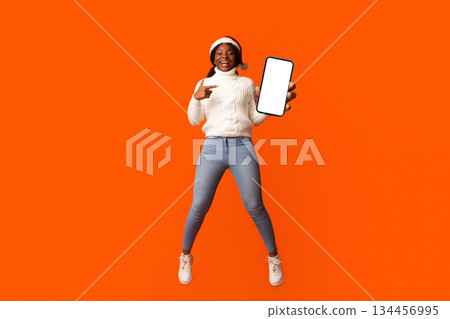 Christmas Offer. Smiling African Lady In Santa Hat Pointing At Smartphone With Black Screen In Her Hand, Demonstrating Free Place For Holiday Mockup Advertisement Or App Design, Orange Background Christmas Offer. Smiling African Lady In Santa Hat Pointing At Smartphone With Black Screen In Her Hand, Demonstrating Free Place For Holiday Mockup Advertisement Or App Design, Orange Background 134456995