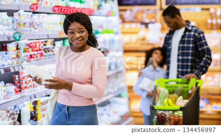 Happy black family with child shopping for products at dairy section of supermarket, copy space. Positive African American parents with kid purchasing groceries at huge mall Happy black family with child shopping for products at dairy section of supermarket, copy space. Positive African American parents with kid purchasing groceries at huge mall 134456997