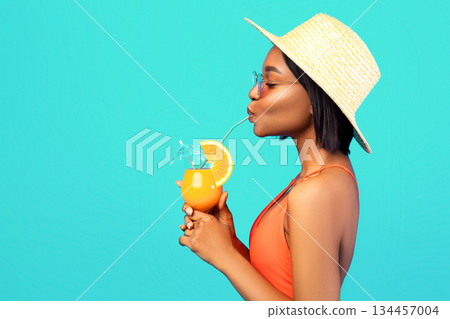 Side view of graceful black woman in swimsuit drinking cool refreshing tropical cocktail on blue studio background. Profile of sensual young lady enjoying tasty exotic drink during beach vacation 134457004