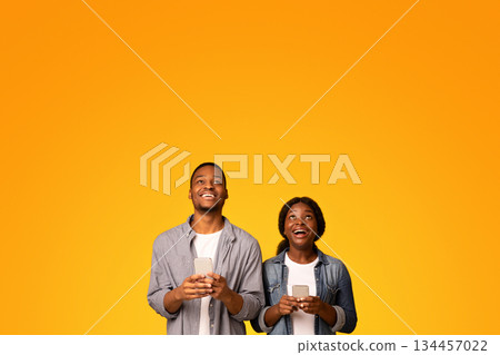 Joyful Black Couple With Smartphones In Hands Looking Up At Copy Space Above Their Heads. Excited African American Man And Woman Standing Isolated Over Yellow Background, Vertical Shot Joyful Black Couple With Smartphones In Hands Looking Up At Copy Space Above Their Heads. Excited African American Man And Woman Standing Isolated Over Yellow Background, Vertical Shot 134457022