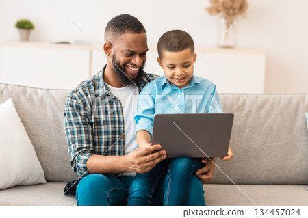 African American Dad Teaching Son How To Use Laptop Computer And Browse Internet Online Sitting On Sofa At Home. Weekend Leisure, Family Gadgets And Technologies Concept 134457024