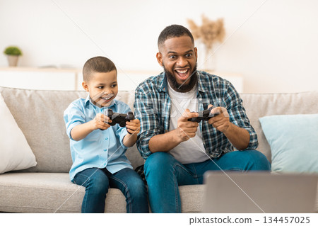 Joyful Black Dad And Son Play Videogame Together Having Fun On Weekend Sitting On Sofa At Home. Young African Daddy Playing Games Spending Time With His Kid Joyful Black Dad And Son Play Videogame Together Having Fun On Weekend Sitting On Sofa At Home. Young African Daddy Playing Games Spending Time With His Kid 134457025