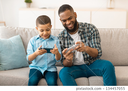 African Father And Son Playing Mobile Games On Smartphone Sitting On Couch At Home. Family Gadgets And Lifestyle, Online Gaming, Application For Phone Concept 134457055