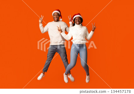 Joyful African American Spouses In Santa Hats Jumping And Showing Peace Gesture, Having Fun Together Over Orange Studio Background, Black Couple Enjoying Celebrating Christmas Holidays, Copy Space Joyful African American Spouses In Santa Hats Jumping And Showing Peace Gesture, Having Fun Together Over Orange Studio Background, Black Couple Enjoying Celebrating Christmas Holidays, Copy Space 134457178