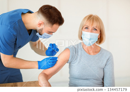 Doctor in medical mask giving covid-19 vaccine shot to senior female patient during immunization campaign at hospital. Mature Caucasian woman getting immunized against viral disease at clinic 134457179