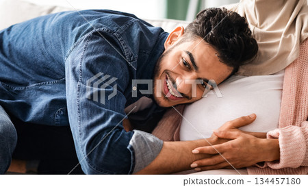 Happy Father. Joyful middle eastern man listening his pregnant wife's tummy, young arab guy tenderly hugging belly of expectant spouse, trying to feel baby kicks, cropped image, closeup 134457180