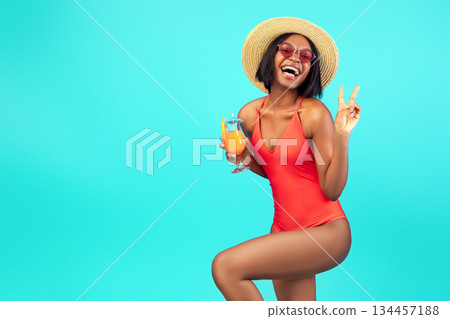 Happy black woman in swimsuit enjoying exotic summer cocktail and showing peace sign at camera, posing over blue studio background. Playful African American lady having fun on beach vacation 134457188