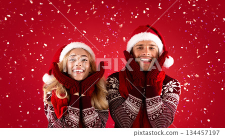 It's Christmas time. Adorable young couple in mittens, Santa hats and scarves on red background 134457197