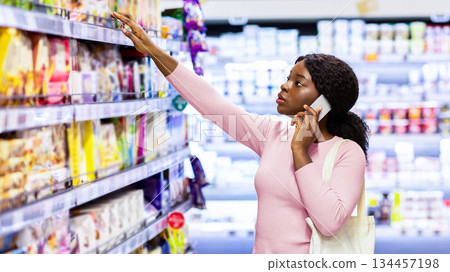 Beautiful young black woman with tote bag making phone call while shopping for food at grocery department of big mall. Lovely African American lady purchasing products at supermarket Beautiful young black woman with tote bag making phone call while shopping for food at grocery department of big mall. Lovely African American lady purchasing products at supermarket 134457198
