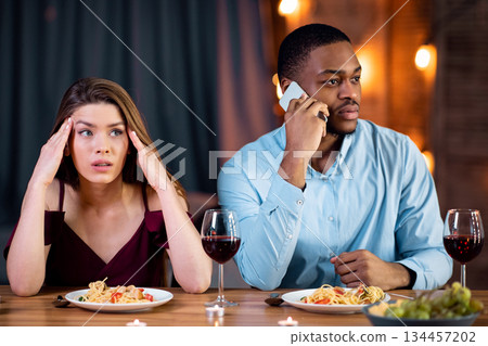 Woman looking unhappy on date while her boyfriend talking on cellphone in restaurant, paying no attention to her and busy using his mobile phone. Multiracial couple dining together at Valentine's day Woman looking unhappy on date while her boyfriend talking on cellphone in restaurant, paying no attention to her and busy using his mobile phone. Multiracial couple dining together at Valentine's day 134457202