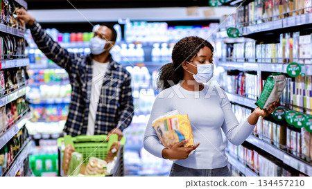Young black woman and man in face masks choosing food products at supermarket during covid quarantine. Millennial couple wearing protection while shopping for groceries at mall Young black woman and man in face masks choosing food products at supermarket during covid quarantine. Millennial couple wearing protection while shopping for groceries at mall 134457210