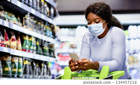 Shopping during covid quarantine concept. Millennial African American woman in protective mask buying food, checking her grocery list on cellphone, purchasing products at modern supermarket Shopping during covid quarantine concept. Millennial African American woman in protective mask buying food, checking her grocery list on cellphone, purchasing products at modern supermarket 134457218