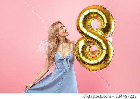 8th March celebration concept. Portrait of lovely blonde lady in dress holding number eight golden balloon for Woman's Day on pink studio background, copy space 8th March celebration concept. Portrait of lovely blonde lady in dress holding number eight golden balloon for Woman's Day on pink studio background, copy space 134457222