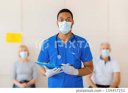 Young African American doctor in antiviral mask and uniform holding clipboard at population immunization centre. Senior patients waiting for covid-19 vaccine injections. Public health concept Young African American doctor in antiviral mask and uniform holding clipboard at population immunization centre. Senior patients waiting for covid-19 vaccine injections. Public health concept 134457251