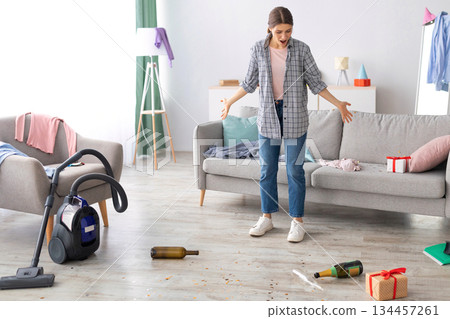 Young woman standing in messy apartment after party, screaming from despair, empty space. Unhappy millennial housewife at dirty room after celebration, rubbish scattered on floor 134457261