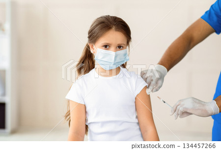 Little girl in medical mask getting covid-19 vaccine injection at health centre. African American doctor immunizing cute kid at clinic. Population protection and infectious disease prevention concept Little girl in medical mask getting covid-19 vaccine injection at health centre. African American doctor immunizing cute kid at clinic. Population protection and infectious disease prevention concept 134457266