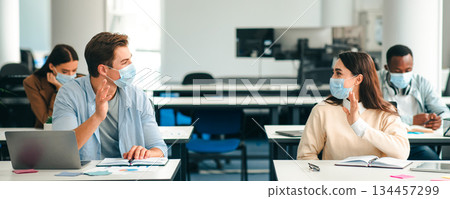 Modern Education Concept. Smiling students wearing protective face masks greeting each other and waving hello at the classroom. Man and woman sitting at desks, studying in high school 134457299