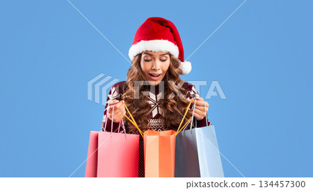 Big Christmas sale. Surprised young lady in Santa hat peeking into gift bags on blue background 134457300