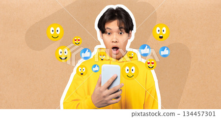 A young Asian guy wearing a yellow hoodie reacts with surprise while chatting on a smartphone. Various emojis surround him, highlighting engagement in social media conversations. 134457301