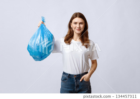 Happy young woman holding blue plastic garbage bag and looking at camera, smiling millennial lady carrying packet with domestic waste while standing on grey studio background, copy space 134457311