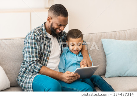 Family Gadgets. Cheerful African Daddy And Little Son Using Digital Tablet Together Watching Cartoons Online And Browsing Internet At Home. Weekend With Father, Technology And Internet 134457314