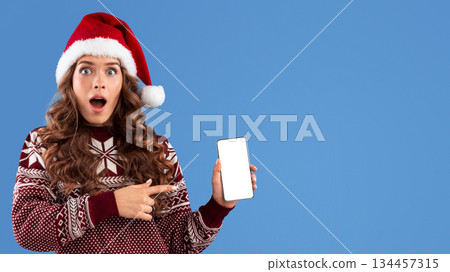Online Christmas shopping. Shocked young girl in Santa hat pointng at mobile phone with empty screen, blue background. Panorama Online Christmas shopping. Shocked young girl in Santa hat pointng at mobile phone with empty screen, blue background. Panorama 134457315