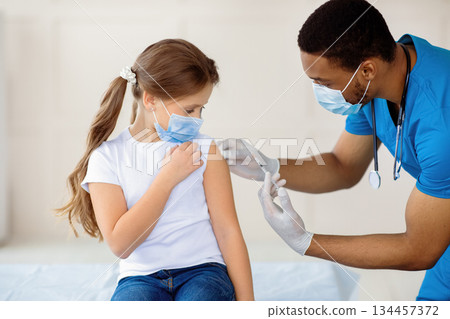 Young black doctor making covid-19 vaccine injection to little girl in face mask at hospital. Child getting coronavirus immunization at clinic. Protection against global virus concept 134457372