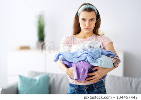 Unhappy millennial housewife holding clothes for washing or ironing, unwilling to do domestic duties, indoors. Portrait of young woman sick and tired of laundry, looking at camera and sulking 134457442