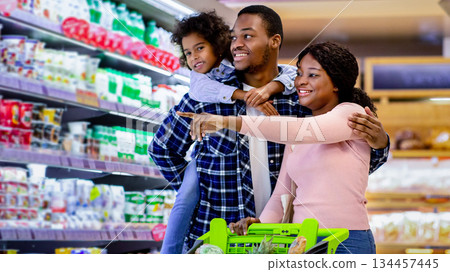 Family shopping. Young black parents with their cute daughter selecting food,pointing at shelves in supermarket. Joyful black girl with mother and father buying products at dairy department of store Family shopping. Young black parents with their cute daughter selecting food,pointing at shelves in supermarket. Joyful black girl with mother and father buying products at dairy department of store 134457445