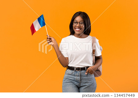 Happy young black woman student in white t-shirt showing french flag, orange studio background, copy space. Smiling african american lady with backpack and glasses showing flag of France 134457449