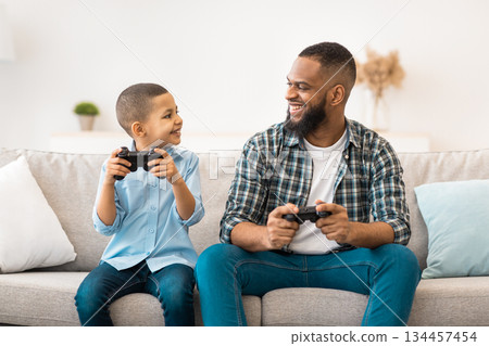 Cheerful African American Father And Son Playing Videogames Holding Game Controllers Having Fun At Home. Weekend Leisure Concept. Young Dad And His Kid Play Computer Games Together Cheerful African American Father And Son Playing Videogames Holding Game Controllers Having Fun At Home. Weekend Leisure Concept. Young Dad And His Kid Play Computer Games Together 134457454