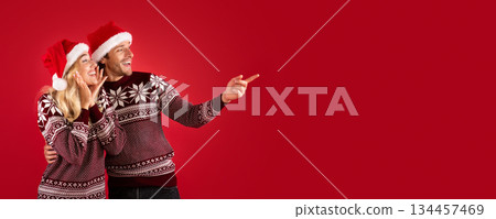 Romantic young couple in Christmas outfits pointing aside at empty space, red background. Panorama Romantic young couple in Christmas outfits pointing aside at empty space, red background. Panorama 134457469