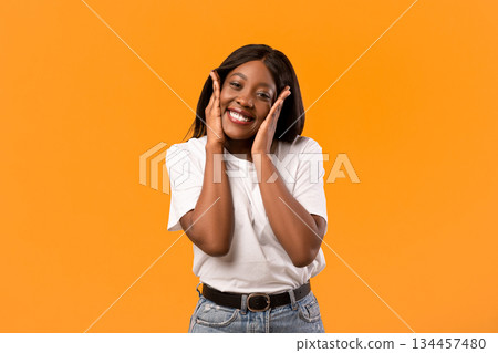 Emotional black lady in white t-shirt smiling and touching her face over orange studio background, happy african american young woman expressing amazement, seeing something exciting, copy space 134457480