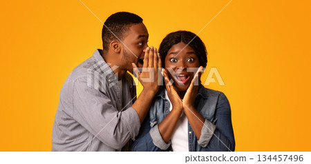 Sharing Secret. Black Guy Gossiping With His Excited Girlfriend, Surprised African American Woman Opening Mouth In Amazement While Standing Together Over Yellow Studio Background, Copy Space 134457496