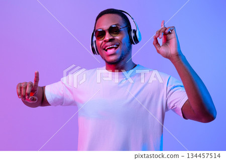 Enjoy The Rhytm. Cheerful African American Guy In Wireless Headphones Listening Music And Dancing, Black Millennial Man Having Fun While Standing Under Neon Lighting Over Purple Background, Copy Space Enjoy The Rhytm. Cheerful African American Guy In Wireless Headphones Listening Music And Dancing, Black Millennial Man Having Fun While Standing Under Neon Lighting Over Purple Background, Copy Space 134457514