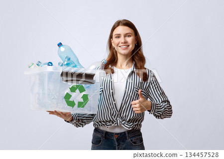 Positive Eco-Friendly Woman Holding Container With Plastic Bottles And Showing Thumb Up Gesture, Smiling Millennial Lady Agitating For Waste Sorting And Recycling Over Gray Studio Background 134457528