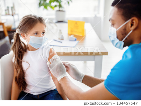 Male doctor or nurse giving shot of coronavirus vaccine to little patient's shoulder at clinic. Lovely girl being immunized against covid-19. Medical service and viral disease prevention concept Male doctor or nurse giving shot of coronavirus vaccine to little patient's shoulder at clinic. Lovely girl being immunized against covid-19. Medical service and viral disease prevention concept 134457547