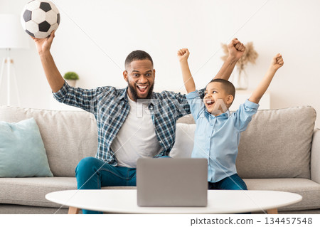 Joyful Black Daddy And Son Watching Sport Game On Laptop Cheering Favorite Soccer Team Sitting On Sofa At Home. Football Fans Watch Online Television And Sports Channels At Computer Concept Joyful Black Daddy And Son Watching Sport Game On Laptop Cheering Favorite Soccer Team Sitting On Sofa At Home. Football Fans Watch Online Television And Sports Channels At Computer Concept 134457548