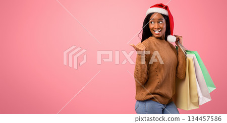 Huge Christmas sale. Attractive young black woman in Santa hat holding gift bags and pointing aside at blank space, pink studio background. African American lady shopping for Xmas Huge Christmas sale. Attractive young black woman in Santa hat holding gift bags and pointing aside at blank space, pink studio background. African American lady shopping for Xmas 134457586