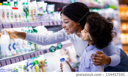 Positive black mom with her child shopping for products at dairy section of mall. Cheerful African American parent with her daughter buying groceries at huge hypermarket Positive black mom with her child shopping for products at dairy section of mall. Cheerful African American parent with her daughter buying groceries at huge hypermarket 134457591