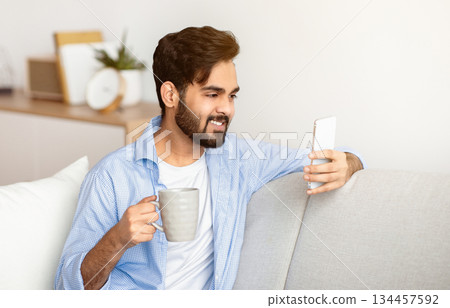 Domestic Leisure. Handsome Eastern Guy Using Smartphone And Drinking Coffee At Home While Resting On Couch In Living Room, Watching Videos Or Making Video Call, Enjoying Weekend Pastime, Free Space Domestic Leisure. Handsome Eastern Guy Using Smartphone And Drinking Coffee At Home While Resting On Couch In Living Room, Watching Videos Or Making Video Call, Enjoying Weekend Pastime, Free Space 134457592