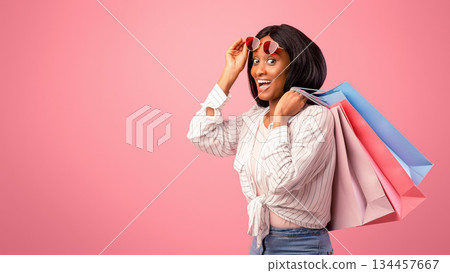 Pretty black woman with shopping bags lifting sunglasses, feeling excited on pink studio background. Lovely African American lady buying holiday gifts, happy over big sale or promo Pretty black woman with shopping bags lifting sunglasses, feeling excited on pink studio background. Lovely African American lady buying holiday gifts, happy over big sale or promo 134457667