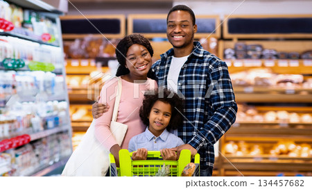 Portrait of smiling black parents with their daughter purchasing groceries at big supermarket. Millennial African American family shopping for products, buying food at hypermarket or mall Portrait of smiling black parents with their daughter purchasing groceries at big supermarket. Millennial African American family shopping for products, buying food at hypermarket or mall 134457682