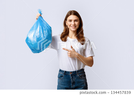 A woman in casual attire holds up a blue plastic bag filled with recyclables, smiling as she encourages others to participate in eco-friendly practices and recycling efforts. A woman in casual attire holds up a blue plastic bag filled with recyclables, smiling as she encourages others to participate in eco-friendly practices and recycling efforts. 134457839