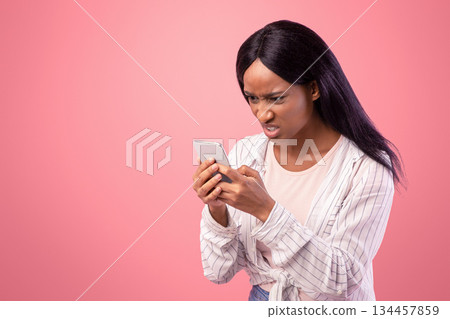 Angry black woman looking at her smartphone with disgust, reading annoying message or news on pink studio background. African American lady feeling stressed at social media publication Angry black woman looking at her smartphone with disgust, reading annoying message or news on pink studio background. African American lady feeling stressed at social media publication 134457859