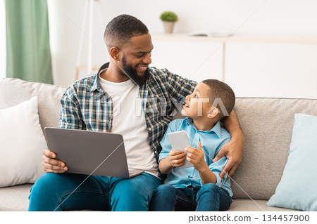 Family Gadgets. Black Daddy And Little Son Using Laptop And Phone Browsing Internet Sitting And Talking Bonding At Home. Weekend With Father, Boy And His Dad Using Computer And Smartphone Together Family Gadgets. Black Daddy And Little Son Using Laptop And Phone Browsing Internet Sitting And Talking Bonding At Home. Weekend With Father, Boy And His Dad Using Computer And Smartphone Together 134457900